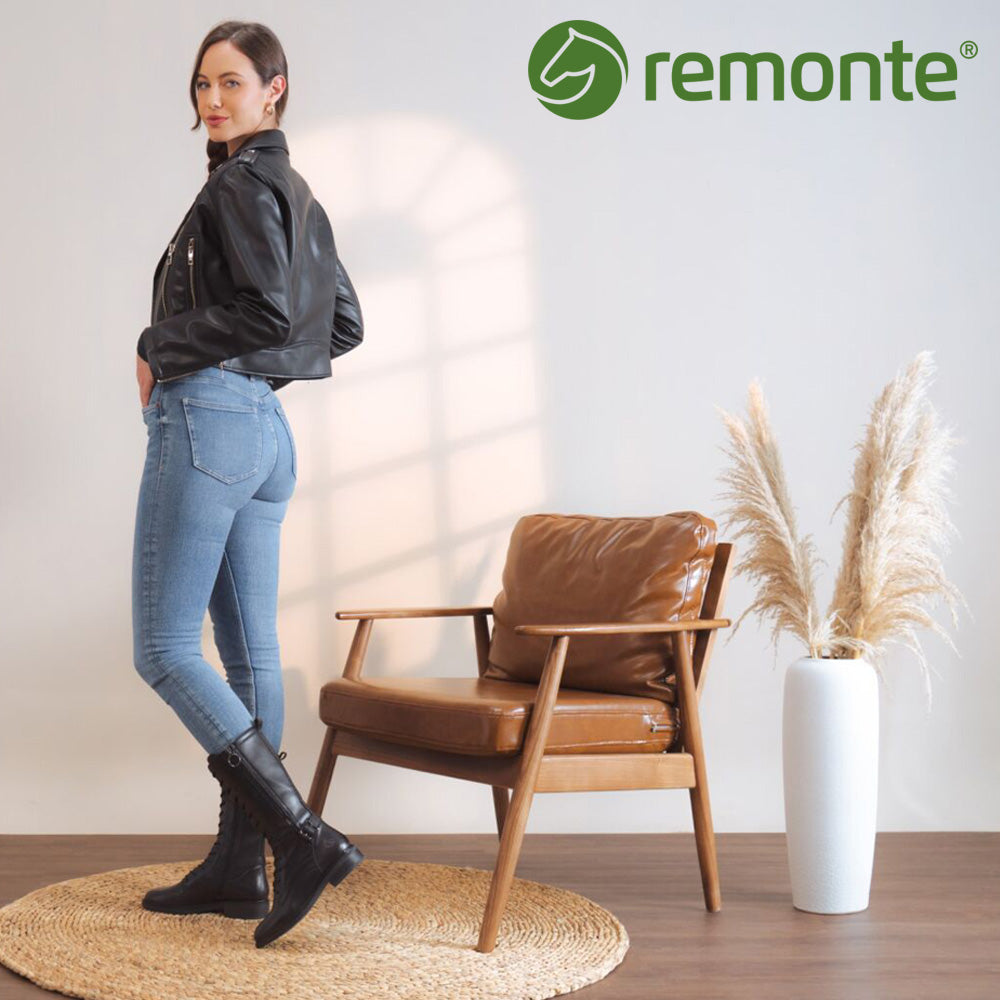 Woman in black jacket and jeans standing next to a brown leather chair with Remonte logo.