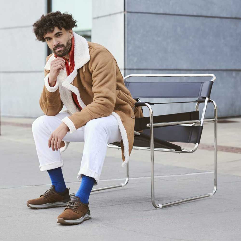 Man sitting on a modern chair wearing a brown coat with white fur trim, white pants, and blue socks.