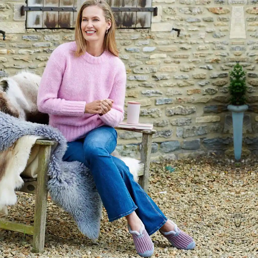 Woman sitting outdoors on a bench wearing a pink sweater and blue jeans, with a stone wall and garden furniture in the background.